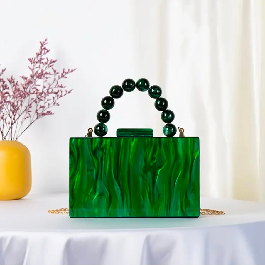 Green marbled handbag with beaded handle on a white surface with a yellow vase and pink branches in the background.