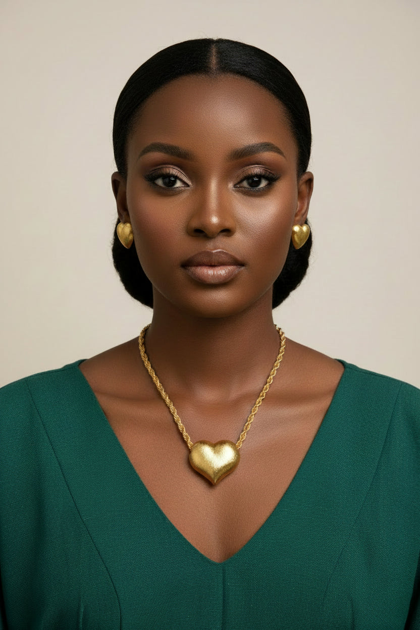 A matte gold jewelry set featuring a large 3D puff heart pendant on a chunky rope chain necklace, on a black female model
