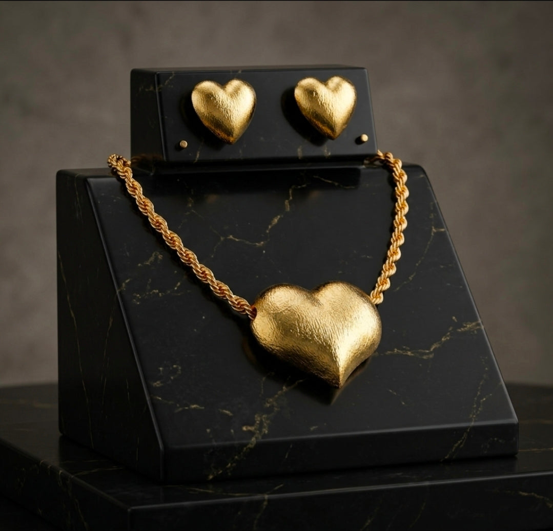 A matte gold jewelry set featuring a large 3D puff heart pendant on a chunky rope chain necklace, displayed alongside matching heart stud earrings on a premium black marbled stand.