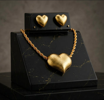 A matte gold jewelry set featuring a large 3D puff heart pendant on a chunky rope chain necklace, displayed alongside matching heart stud earrings on a premium black marbled stand.
