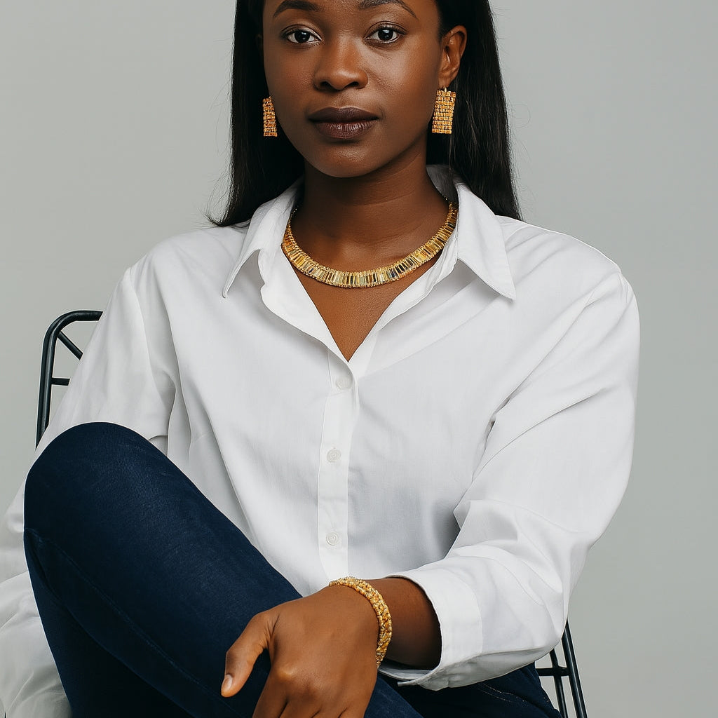Woman wearing a white shirt, gold necklace, and gold bracelet sitting on a chair against a gray background