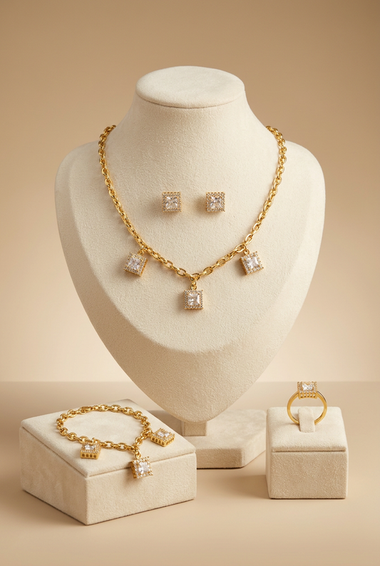 A 4-piece gold-plated jewelry set displayed on a brown mannequin bust, featuring a matching necklace, link bracelet, stud earrings, and an adjustable ring, all adorned with square-cut crystal charms surrounded by a pavé border.