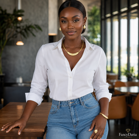 A woman wearing a white button-down shirt, blue jeans, a gold necklace, and a gold bracelet, posing in a modern indoor setting with a wooden table in the foreground.